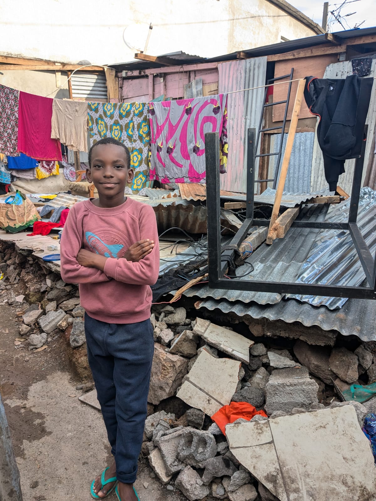 Un petit garçon pose devant sa maison de tôle détruite, tout est à terre, des draps sont étendus sur une corde à linge.