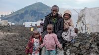 Ladislas entouré de ses enfants dans le camp de déplacés de Bulengo, en septembre 2024. Un homme souriant est entouré de trois enfants. Ils sont assis sur un muret en pierre noir. Derrière eux, on devine des alignements de tentes et, au loin, une colline verdoyante.