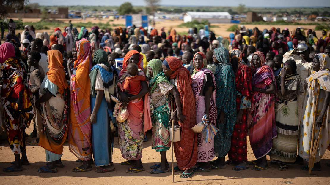 Women stand one behind the other, in a line that can be seen continuing far behind them. Each is holding an empty basin or container.