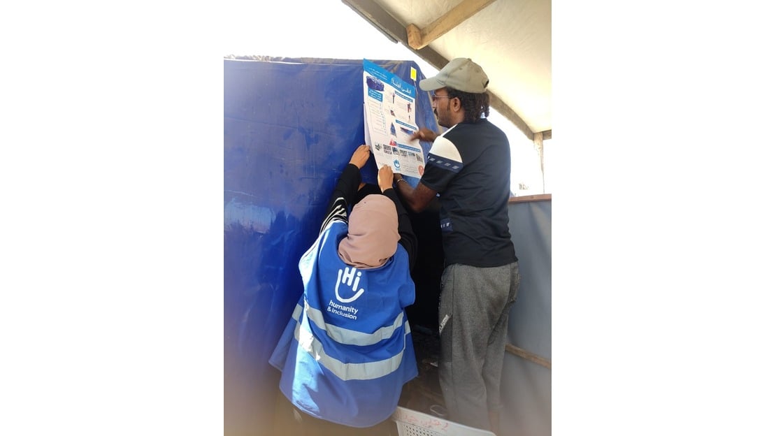 Une femme staff HI, qui porte un gilet bleu avec le logo HI, et un homme, sont en train d'installer une pancarte d'information sur les restes explosifs. Ils sont dans un camp de réfugiés, ils collent l'affiche sur un mur. 